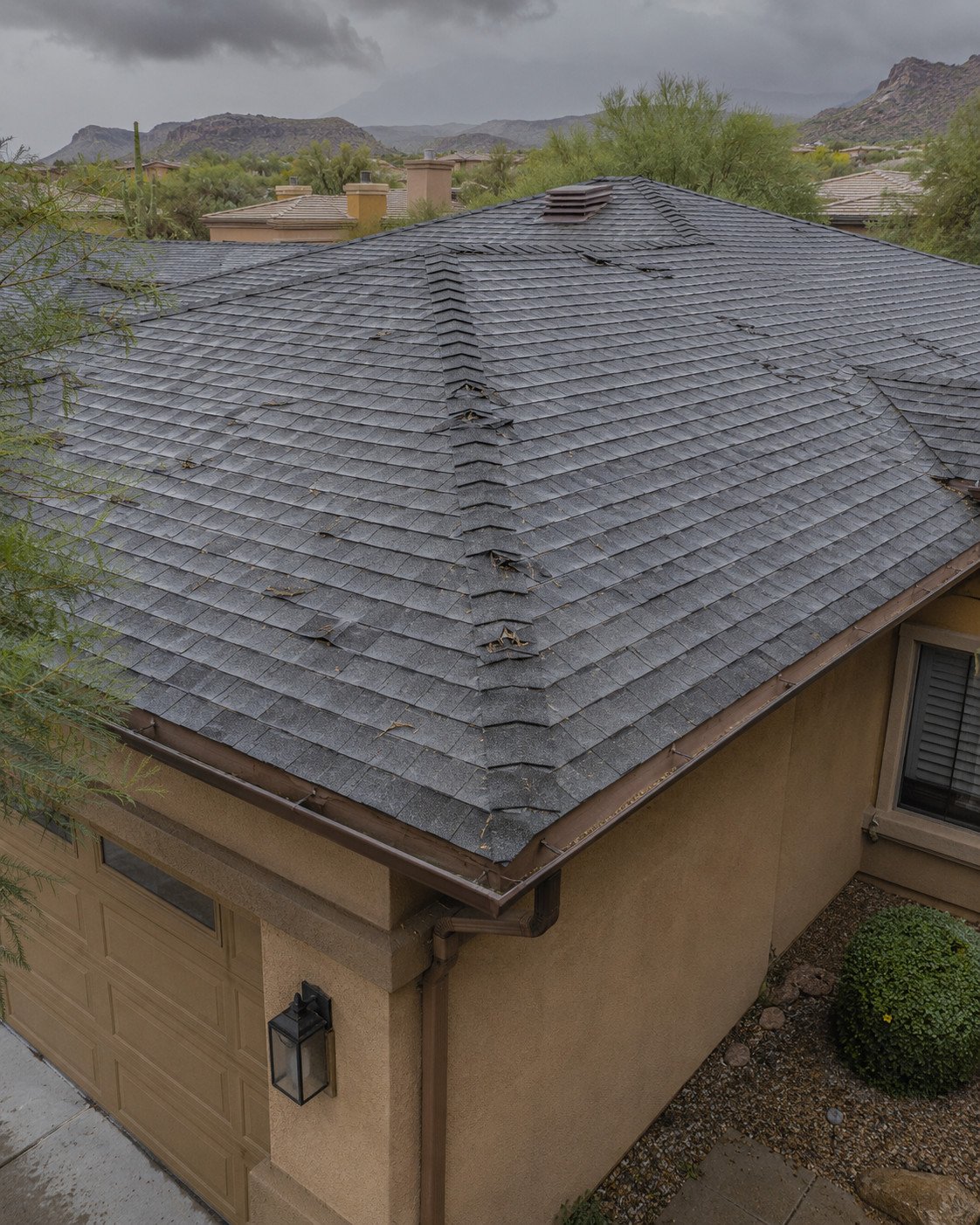 Architectural shingle roof under a monsoon sky with hip shingles creased, lifted and torn by hail and wind